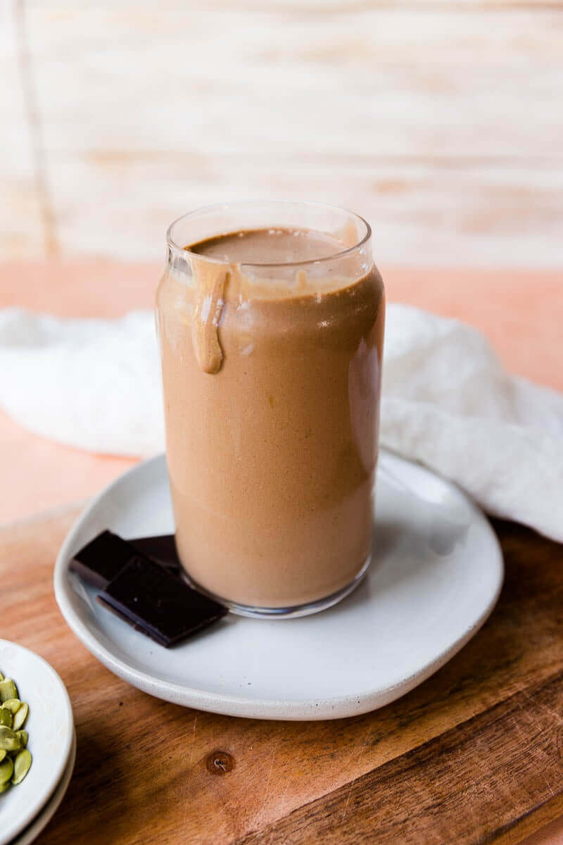 A tall glass filled with a thick chocolate smoothie, served on a white plate with a few pieces of dark chocolate on the side, placed on a wooden surface.