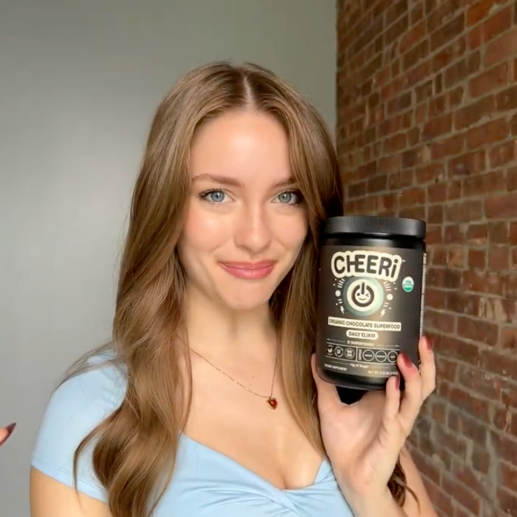 Woman holding a container of 'Cheer!' supplement against a brick wall.